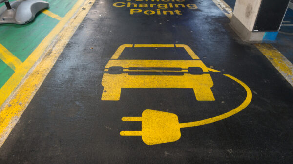 Line markings in a car parking bay say Electric Vehicle Charging Point in yellow letters on black background.An image of a car and charging lead are also visible. Image