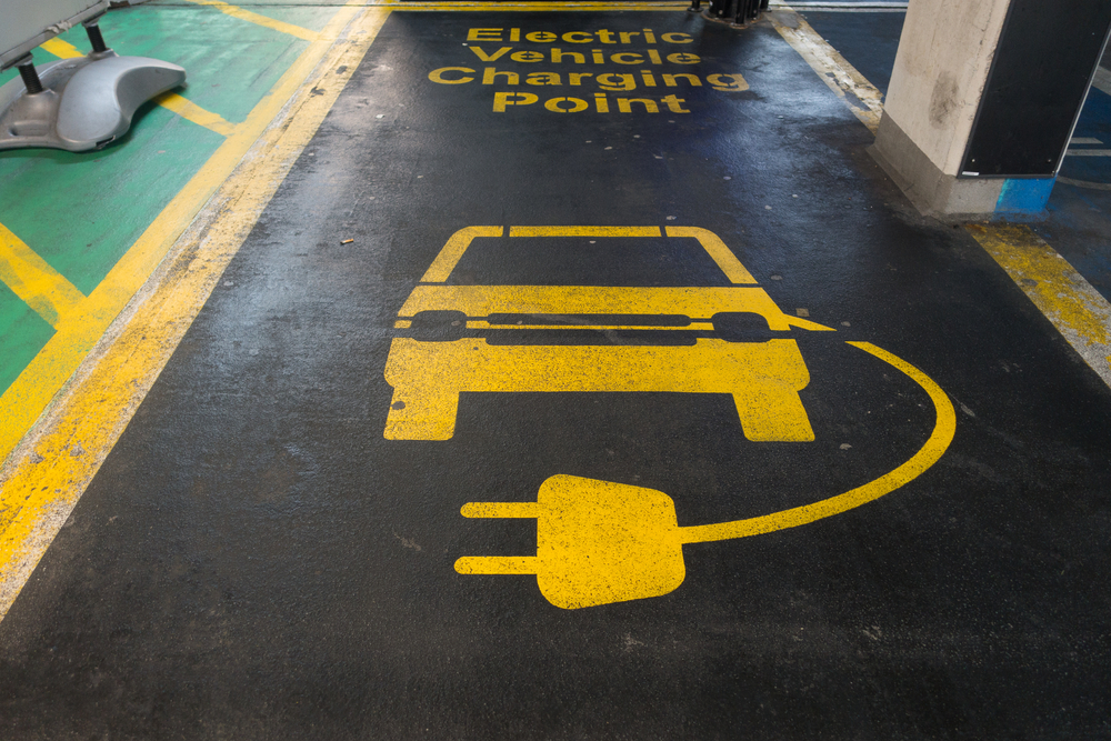 Line markings in a car parking bay say Electric Vehicle Charging Point in yellow letters on black background.An image of a car and charging lead are also visible. Image