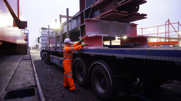 Bolton, Greater Manchester / UK Driver Wearing Full PPE and Chaining Down a Load of Steel Structures onto a Semi Truck Flat bed with Metal Posts Fitted. manufacturing
